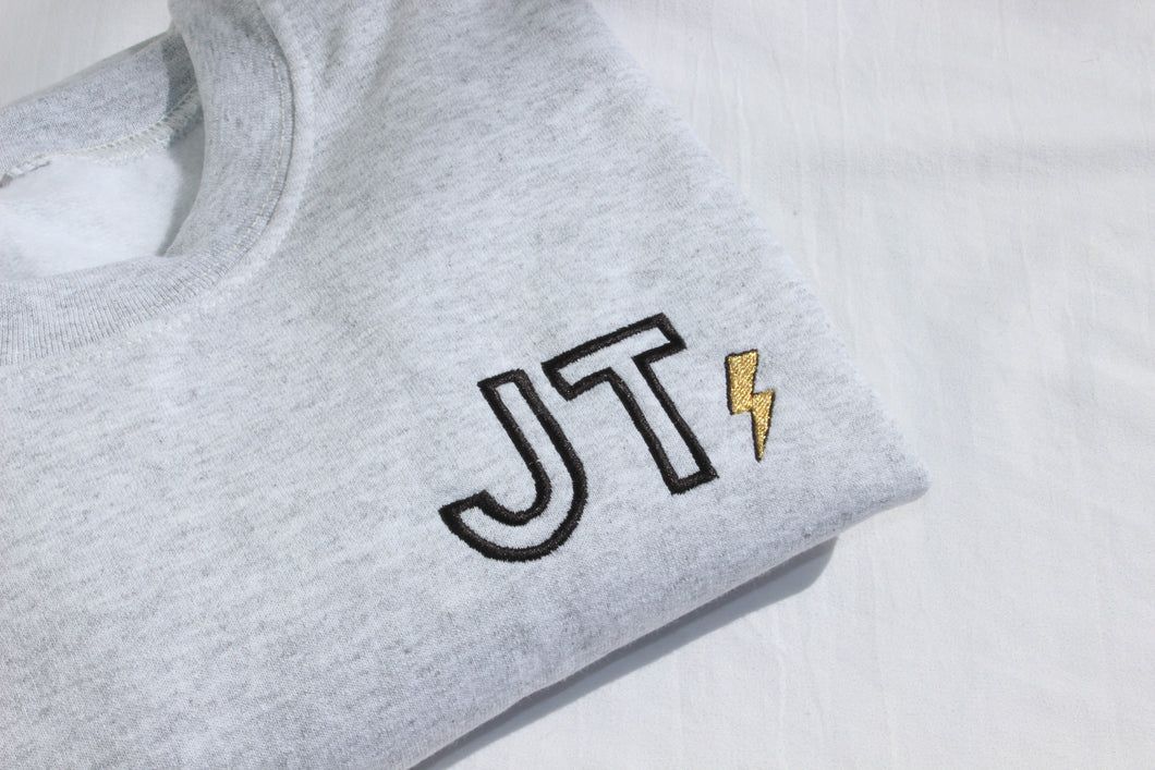 Ash grey crewneck with initials shown in Outline font with a metallic lightning bolt on the side of the initials.